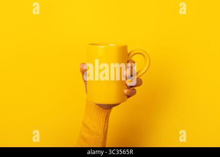Hand with yellow nails and sweater holding a plain yellow ceramic mug in front of a solid yellow background. Stock Photo