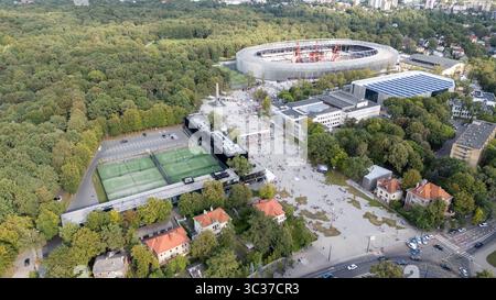Aerial view of a sports complex surrounded by lush greenery, featuring a large stadium, tennis courts, and nearby buildings. People are gathered in th Stock Photo