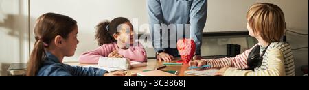 Header of group of diverse children sitting at table listening to Caucasian male teacher explaining lesson, school supplies scattered on desk, students showing attentive expressions Stock Photo