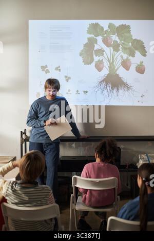 Caucasian middle aged man teaching group of diverse children about plant biology and strawberry growing, standing in front of projected botanical diagram, students sitting and listening attentively Stock Photo