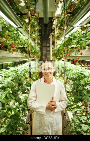 Portrait of young Caucasian woman scientist standing in vertical farm, holding clipboard and pen, smiling at camera, surrounded by rows of strawberry plants in hydroponic greenhouse Stock Photo
