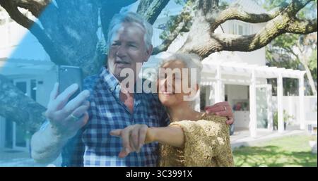 Taking selfie, mature couple standing under gnarled tree in backyard, with smartphone and pergola Stock Photo