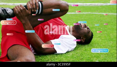 Clutching right knee while lying on grass field wearing red jersey, striped socks and digital tags Stock Photo