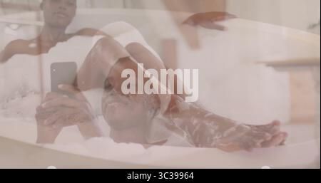 Reclining woman in towel wrap holding smartphone in bathroom, with standalone tub foam bubbles Stock Photo