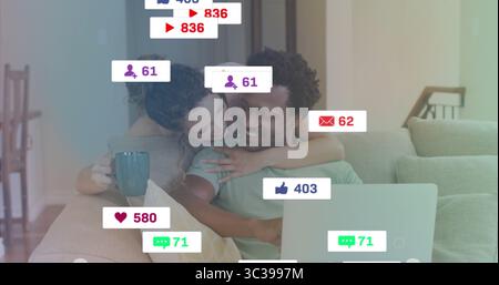 Smiling couple hugging on couch, using laptop browsing with floating icons, holding teal coffee mug Stock Photo