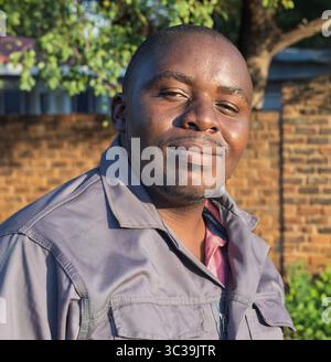 Smiling single African overweight man in workwear , outdoors with a brick wall background, standing, expressing happiness, bright natural sunlight, ca Stock Photo