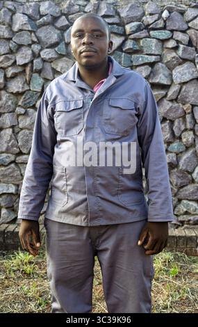 Smiling single African overweight man in workwear , outdoors with a brick wall background, standing, expressing happiness, bright natural sunlight, ca Stock Photo