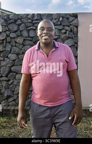 A cheerful single African man standing outdoors in front of a wall made of stacked stones, wearing a casual pink polo shirt and gray pants, smiling wa Stock Photo