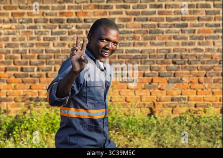 Young African man in blue workwear, orange stripe, walking on the street smiling and making a peace sign, standing outdoors in front of a brick wall w Stock Photo