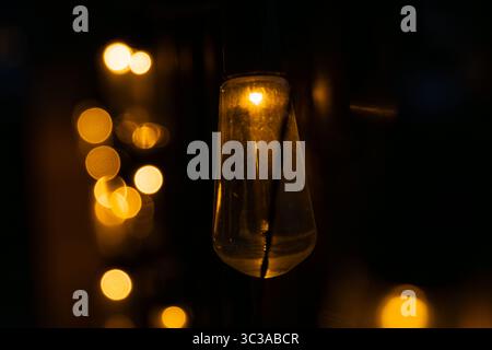Glowing light bulb hanging in focus, with other lights creating a warm bokeh effect in a dark background Stock Photo