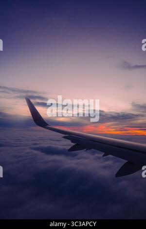 View from airplane window showing aircraft wing above fluffy clouds during colorful sunset or sunrise Stock Photo