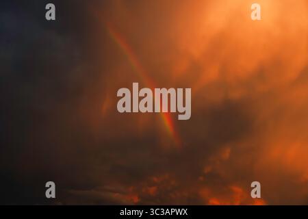 Dramatic sky with a vivid rainbow emerging through fiery orange storm clouds during sunset Stock Photo