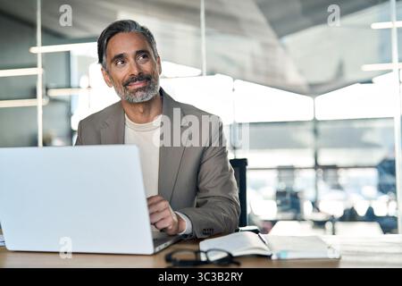 Smiling middle aged business man sitting at desk using laptop looking away. Stock Photo
