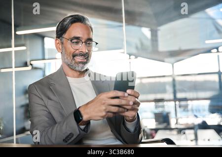Smiling mature business man executive sitting at desk using smartphone. Stock Photo