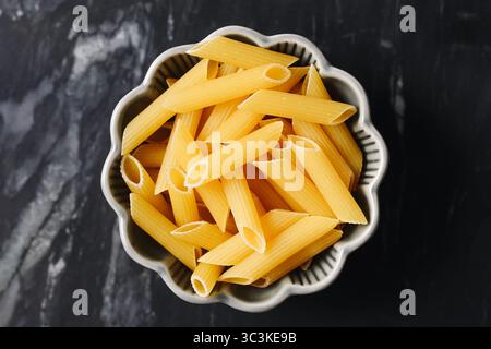 Penne pasta in ceramic bowl. Uncooked yellow noodles on black background. Italian food ingredient top view. Culinary concept for cooking traditional. Stock Photo