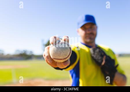 White baseball being held toward camera with black glove resting on baseball field under blue sky Stock Photo