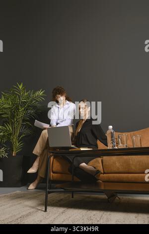 Two businesswomen sitting on an office couch. One is reviewing documents, while the other focuses on a laptop. A green plant adds a fresh touch to the Stock Photo