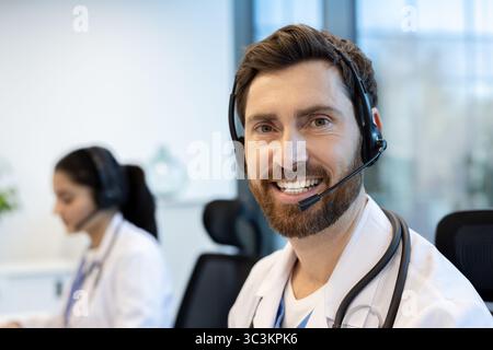 A doctor wearing a headset smiles at the camera while a colleague is in the background. The image shows a medical professional in an office setting. Stock Photo