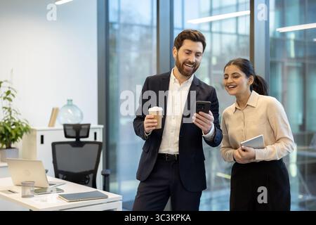 Two diverse business colleagues share a laugh while looking at a smartphone in a modern office environment. Stock Photo