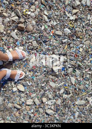 A person is standing on a beach with trash all around them. The beach is littered with plastic bottles, and the person's feet are covered in debris Stock Photo