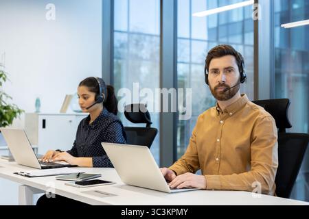 Two call center employees are working at their desks, each wearing a headset and using a laptop. The man looks directly into the camera. Stock Photo