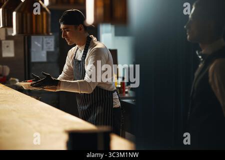 Young adult Caucasian man wearing apron and cap serving food at restaurant counter, extending hands with plate toward customer, working in professional kitchen environment Stock Photo