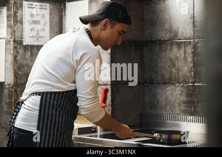 Young adult Caucasian man wearing apron standing in professional kitchen cooking food in frying pan on stovetop, concentrating on preparing meal in restaurant setting Stock Photo