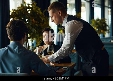 Caucasian young adult man serving wine to Caucasian young adult man and Caucasian young adult woman sitting at restaurant table, waiter placing glasses while couple waiting Stock Photo