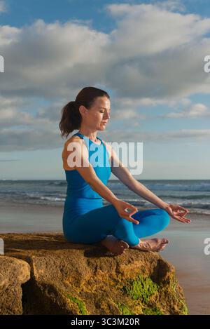 sporty woman sitting on a rock meditating and practicing yoga at the beach Stock Photo