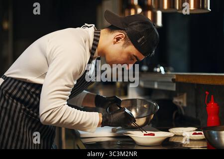Young adult Caucasian man wearing apron and cap preparing dessert in professional kitchen, using tweezers to carefully arrange garnish on plate, focused on culinary presentation Stock Photo