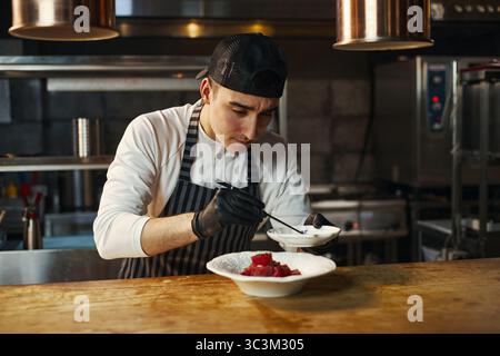 Young adult Caucasian man wearing chef uniform preparing gourmet dish in professional restaurant kitchen, using tweezers to arrange food on plate, focused on presentation Stock Photo