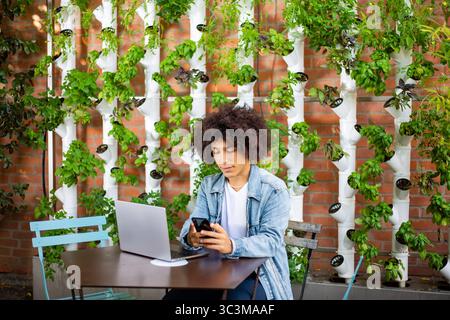 Serious, focused ethnic men using laptop, smartphone at outdoor internet cafe eco friendly area with hydroponic systems. Free green open space place Stock Photo