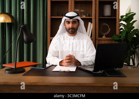 Middle-eastern businessman wearing emirati kandura working in the office - emirati businessman in traditional kandura working on laptop and digital ta Stock Photo