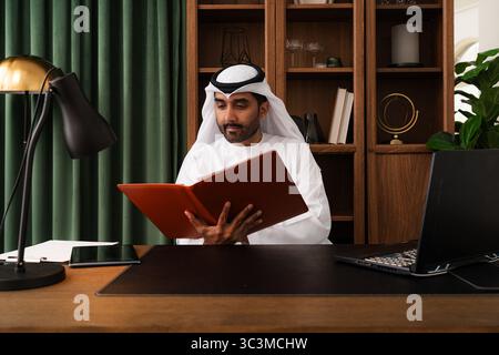 Middle-eastern businessman wearing emirati kandura working in the office - focused arab entrepreneur in traditional kandura analyzing paperwork at an Stock Photo