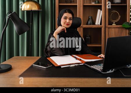 Arab female doctor wearing a black hijab and abaya sits at her desk in a modern office, working on her laptop with a notebook and pen, exuding profess Stock Photo