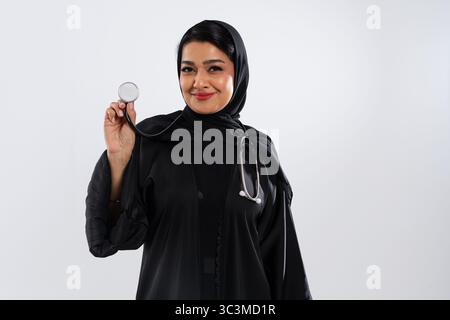 Smiling arab female doctor wearing black hijab and abaya holding a stethoscope while posing in a studio setting with a white background, representing Stock Photo