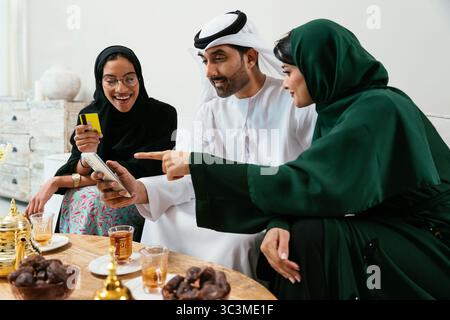 Emirati family enjoying quality time at home, sharing tea and dates, while engaging in online shopping with a mobile phone and credit card, embodying Stock Photo