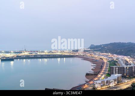 Dover town seen from the hill top castle during evening blue hour. Seafront and beach leading to the Western docks and port area illuminated at night. Stock Photo