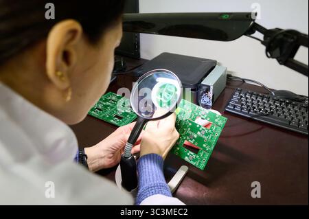 Expert technician examines circuit boards under magnifying glass in a modern electronics workshop Stock Photo