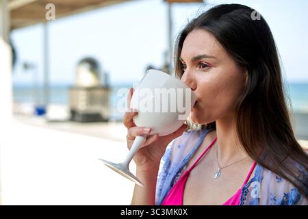 A young woman of Caucasian descent sips a large beverage, standing by the beach under a bright sun. The atmosphere exudes relaxation and summer vibes Stock Photo