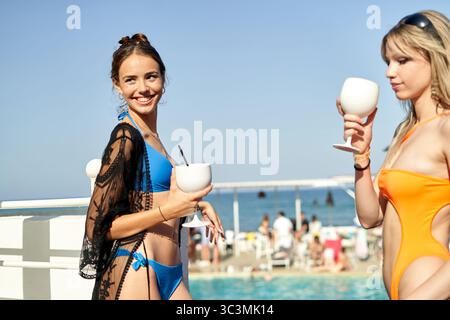 Bright sunlight shines on two young women by the poolside, enjoying tropical drinks while surrounded by vacationers. The beach is lively, enhancing th Stock Photo