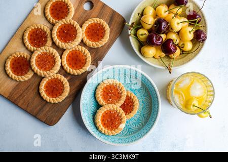 Delicious homemade cookies topped with sweet marmalade, accompanied by fresh white and dark sweet cherries. Perfectly styled on a wooden board and cer Stock Photo