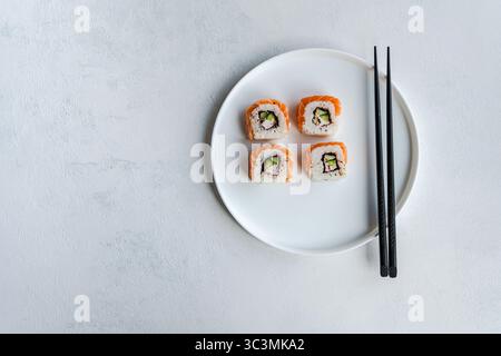 Top view of California roll with salmon on a white ceramic plate placed on a light white textured background. Accompanied by black chopsticks, highlig Stock Photo