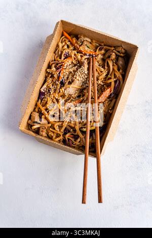 Top view of a delicious handmade Asian noodle dish with chicken, carrots, and red cabbage. Garnished with sesame seeds and served in a brown paper box Stock Photo