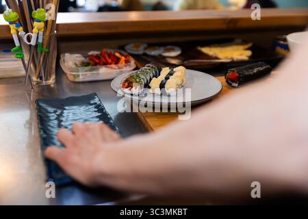 A close-up view of sushi preparation at a restaurant counter. A plate with assorted sushi rolls and utensils nearby creates an inviting Japanese culin Stock Photo
