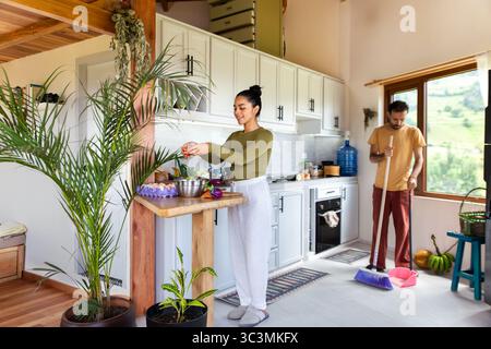 A couple spends leisurely time at home, engaged in daily activities. One partner cooks vegetables in the kitchen, while the other sweeps the floor, sh Stock Photo