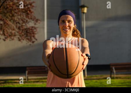A woman stands outdoors holding a basketball, ready to play in a street setting. She wears a headband and athletic wear, illuminated by the warm glow Stock Photo