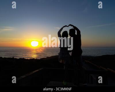 Young couple in love sitting in front of spectacular sunset view over the sea and doing heart with their arms together. Love, relationship concept. Stock Photo