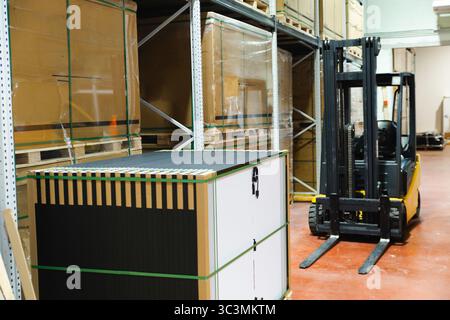 A stack of solar panels is stored in a well organized warehouse, ready for distribution. A yellow forklift is positioned nearby, highlighting the effi Stock Photo