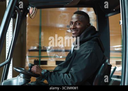 A man smiles while operating a forklift in a solar panel warehouse. Surrounded by boxes, he demonstrates efficiency in managing warehouse logistics an Stock Photo